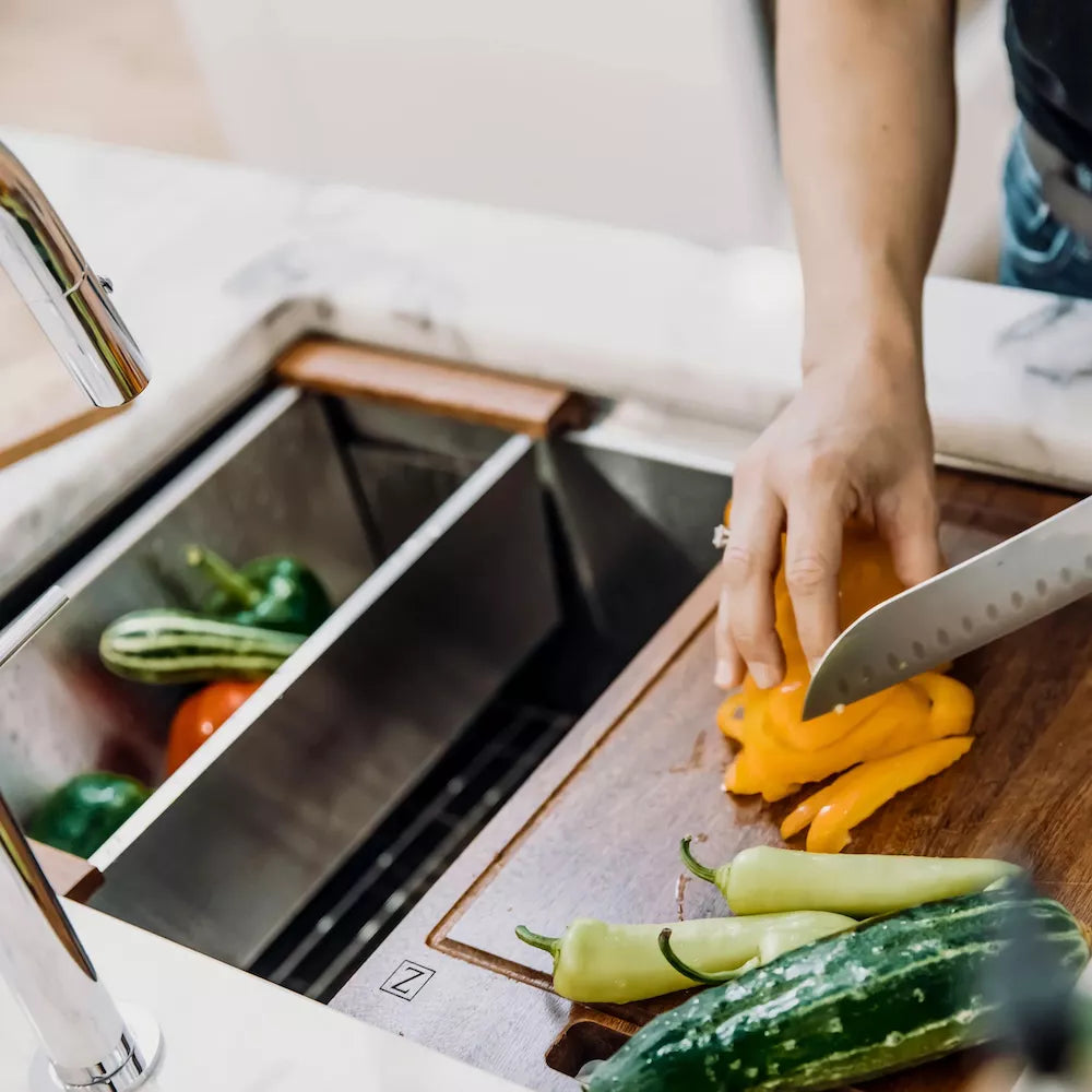 A close-up lifestyle shot of a person slicing a yellow bell pepper on a wooden cutting board next to a double-bowl stainless steel sink filled with vegetables.