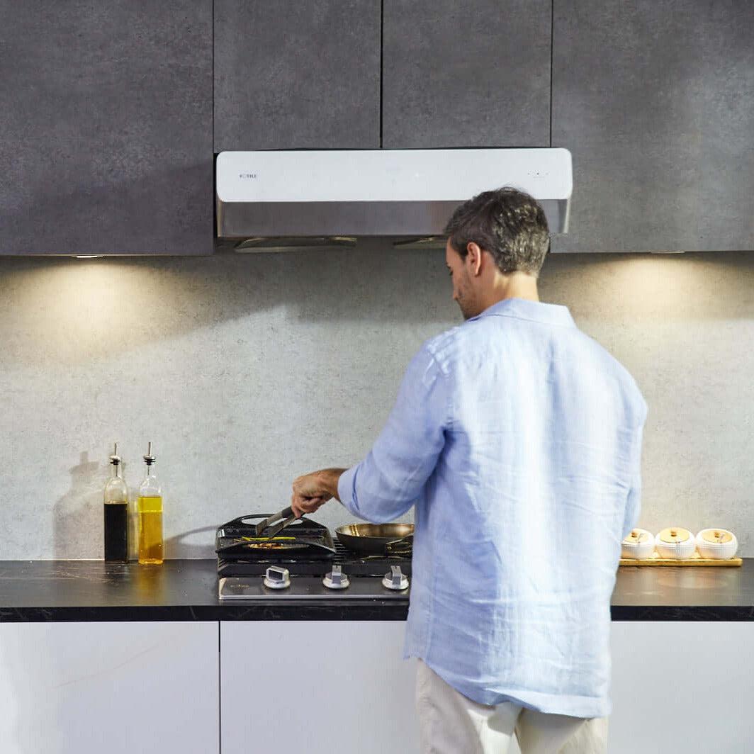 Sleek modern kitchen corner showcasing a Fotile Pixie Air Series Slim Line under cabinet range hood above a gas cooktop, with a minimalist coffee maker, white cups on a wooden holder, and small candles adding a homey touch to the monochromatic decor.