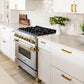 ZLINE Autograph Edition 36 in. 4.6 cu. ft. Legacy Dual Fuel Range with 6 Burner Gas Cooktop and Electric Convection Oven in Stainless Steel and Polished Gold Accents (RAZ-36-G) in a kitchen with white cabinetry, gold handles, and greenery.