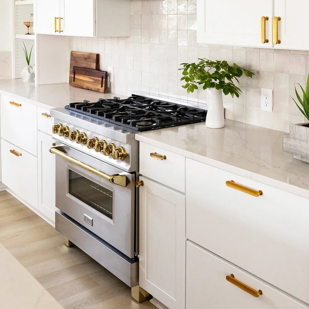 ZLINE Autograph Edition 36 in. 4.6 cu. ft. Legacy Dual Fuel Range with 6 Burner Gas Cooktop and Electric Convection Oven in Stainless Steel and Polished Gold Accents (RAZ-36-G) in a kitchen with white cabinetry, gold handles, and greenery.