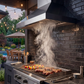 An outdoor kitchen setup with a VICTORY Twister 1200 CFM Outdoor Grill Range Hood, charcoal grill, and steaks cooking on a dark brick wall, with a patio dining area and pool visible in the background.