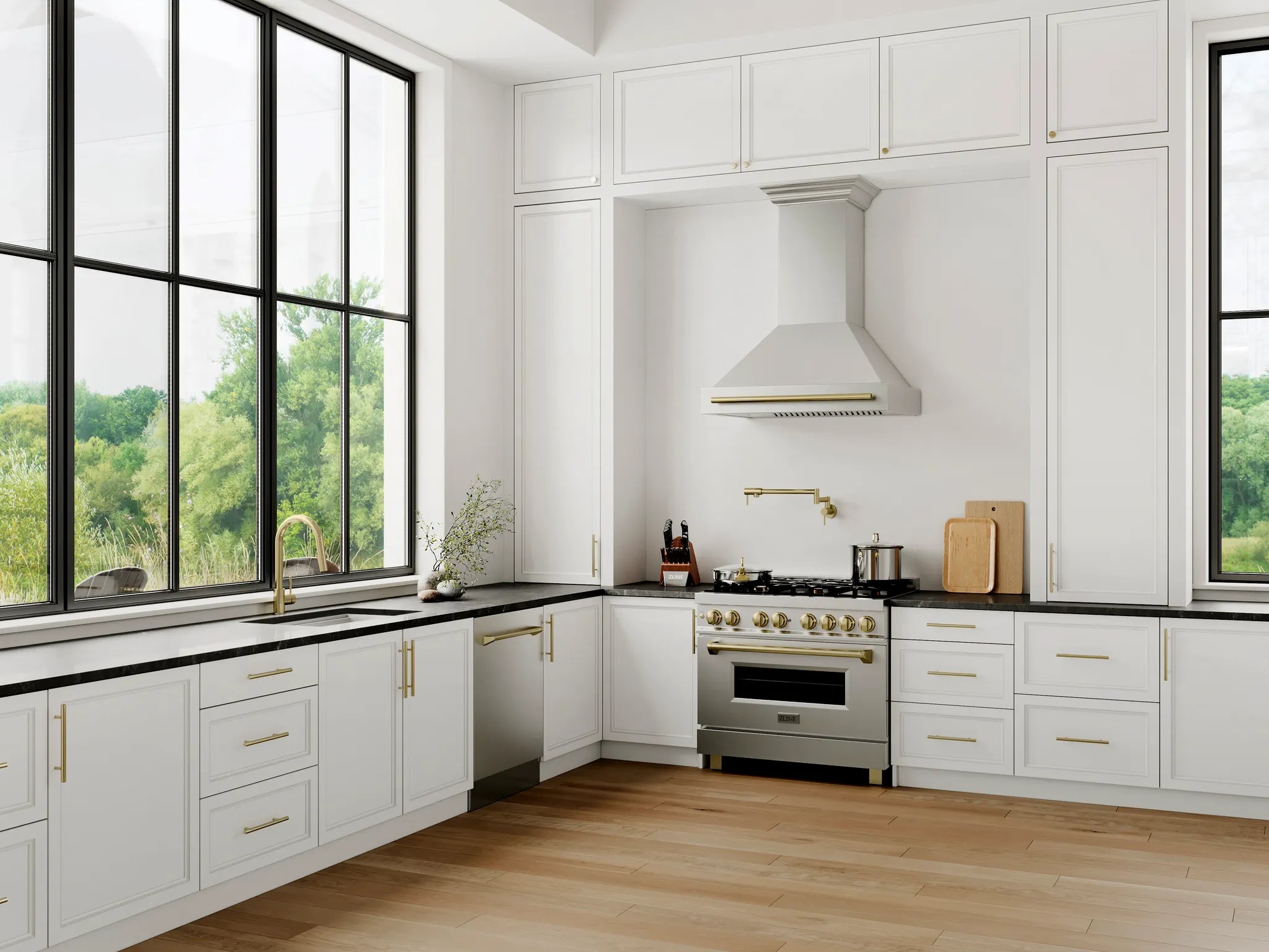 A modern farmhouse kitchen featuring white shaker-style cabinets with gold handles, black countertops, a stainless steel range hood and gas stove, and light wood flooring. Large black-framed windows on both sides offer a view of green trees.