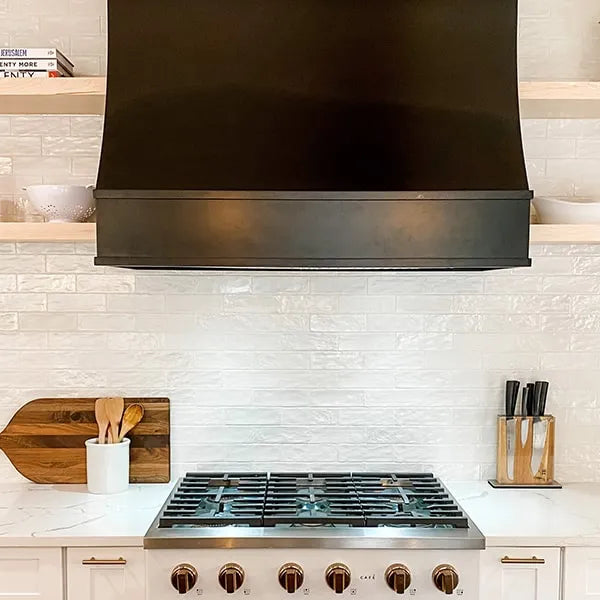 A kitchen scene featuring a sloped black range hood, set into white cabinetry with a white subway tile backsplash.
