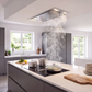A modern kitchen island with a VICTORY Horizon 42 in. 900 CFM Flush Ceiling Mount Range Hood in Stainless Steel. The kitchen features gray cabinetry, a white countertop, cooking pots, stacked plates, and a wooden cutting board.