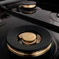 ZLINE Autograph Edition 60 in. 7.4 cu. ft. Legacy Dual Fuel Range with 9 Burner Gas Cooktop and 2 Electric Convection Ovens in Stainless Steel with White Matte Doors and Champagne Bronze Accents (RAZ-WM-60-CB) close-up, brass burners on black porcelain cooktop with no grates.