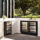 ZLINE Autograph Edition 24 in. Touchstone Dual Zone 44 Bottle Wine Cooler With Black Matte Glass Door And Polished Gold Handle (RWDOZ-BLM-24-G) side, in an outdoor patio area against a stone wall and greenery.