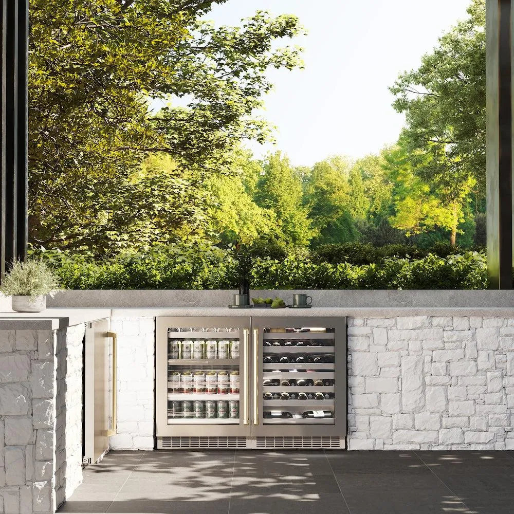 ZLINE Autograph Edition 24 in. Touchstone 151 Can Beverage Fridge with Stainless Steel Glass Door and Champagne Bronze Handle (RBSOZ-GS-24-CB) in an outdoor kitchen with stone walls and greenery