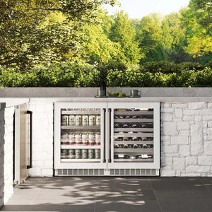Outdoor kitchen with a ZLINE Autograph Edition 24 in. Touchstone Dual Zone 44 Bottle Wine Cooler with White Matte Glass Door and Matte Black Handle (RWDOZ-WM-24-MB) against a stone wall and greenery.