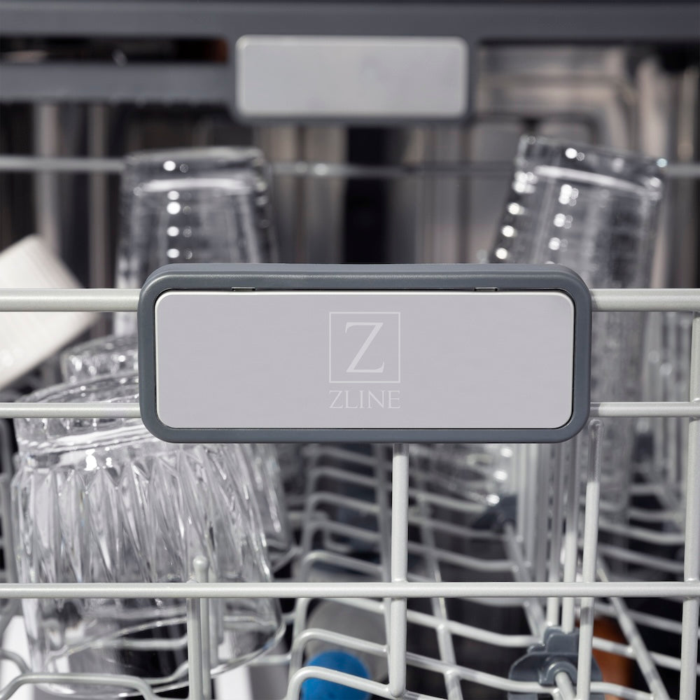 ZLINE Autograph Edition 24 in. Monument Series 3rd Rack Top Touch Control Tall Tub Dishwasher in Stainless Steel with Polished Gold Helix Handle, 45dBa (DWMTZ-30424-KG) logo on dish rack with dishes in the background.