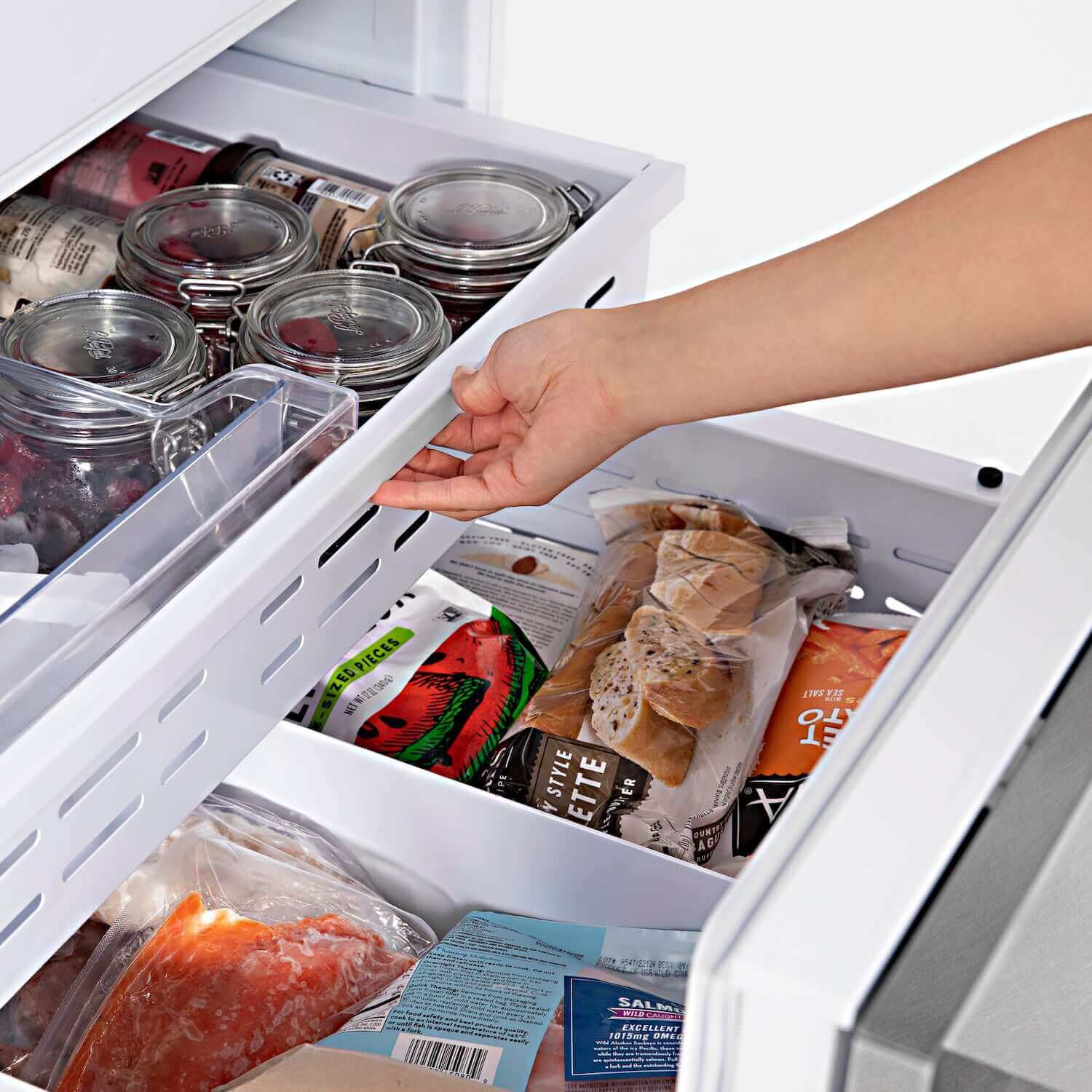 Person opening bottom freezer compartments on built-in refrigerator with food inside.