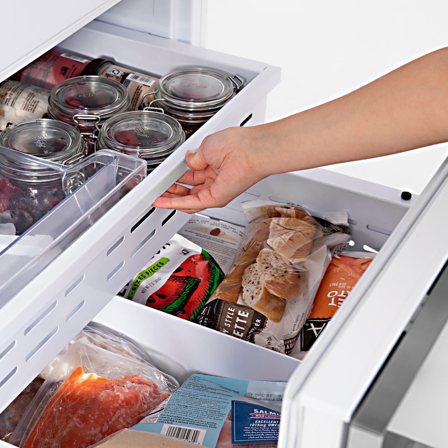 Person opening bottom freezer compartments on built-in refrigerator with food inside.