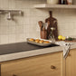 A kitchen scene with beige square wall tiles, a light wood countertop, a ZLINE 36 in. Induction Cooktop with Anti-Scratch Glass and 5 Elements (FCIND-36), wooden cabinets, and kitchen accessories including a baking dish with food, utensils, lemons, and a cloth.