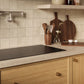 A modern kitchen scene with a ZLINE 36 in. Induction Cooktop with Anti-Scratch Glass and 5 Elements (FCIND-36), beige tiled backsplash, light wood cabinets, and dark wood cutting boards and utensils on the counter.