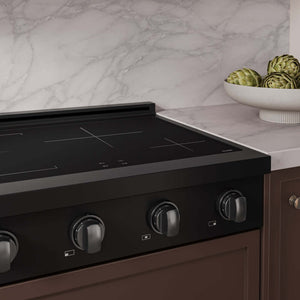 A close-up of a ZLINE Autograph Edition 30 in. Paramount Induction Rangetop with 4 Element Cooktop in Black Stainless Steel and Polished Gold Accents (SINRTBZ-30-G) set into a brown cabinetry base against a white marble backsplash. A white bowl of artichokes sits on the marble countertop to the right.