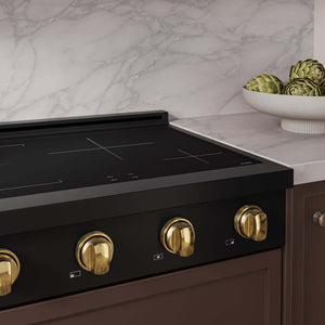 A close-up of a ZLINE Autograph Edition 30 in. Paramount Induction Rangetop with 4 Element Cooktop in Black Stainless Steel and Polished Gold Accents (SINRTBZ-30-G) set into a brown cabinetry base against a white marble backsplash. A white bowl of artichokes sits on the marble countertop to the right.