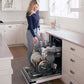 A woman stands in a bright white kitchen next to an open ZLINE Autograph Edition 24 in. Tallac Series 3rd Rack Dishwasher with Auto Door Open Drying, 51dBa, Panel Ready with Matte Black Handle (DWVZ-24-MB) loading plates and glassware into the lower rack.