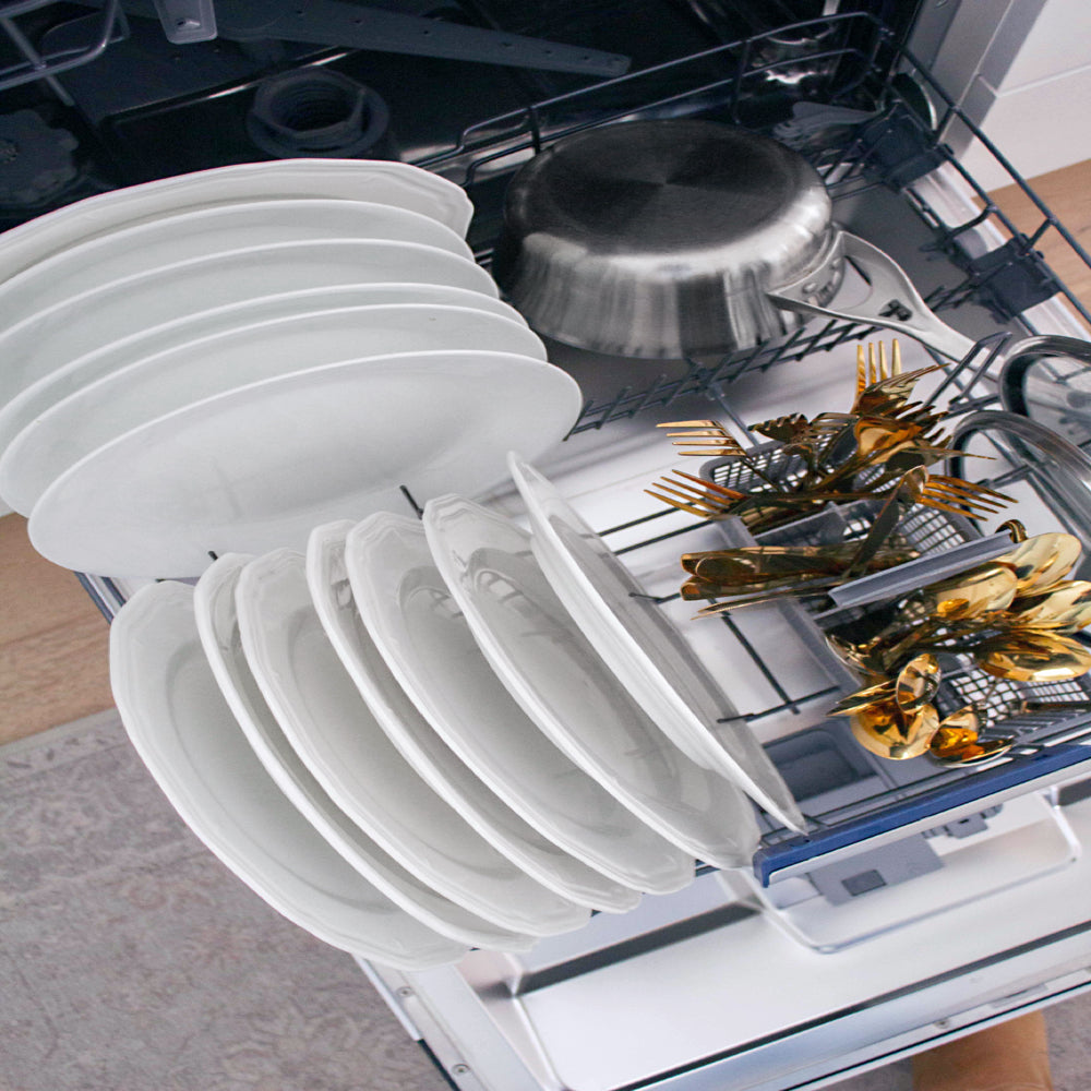 A woman stands in a bright white kitchen next to an open ZLINE Autograph Edition 24 in. Tallac Series 3rd Rack Dishwasher with Auto Door Open Drying, 51dBa, Panel Ready with Matte Black Handle (DWVZ-24-MB) loading plates and glassware into the lower rack.