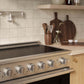 A ZLINE 36 in. 5.2 cu. ft. Marino Induction Range with 5 Element Cooktop and Electric Convection Oven in Stainless Steel (MINR36), set into a light wood kitchen cabinet. The background features beige square tiles, a wooden shelf, and dark wood cutting boards on the countertop.