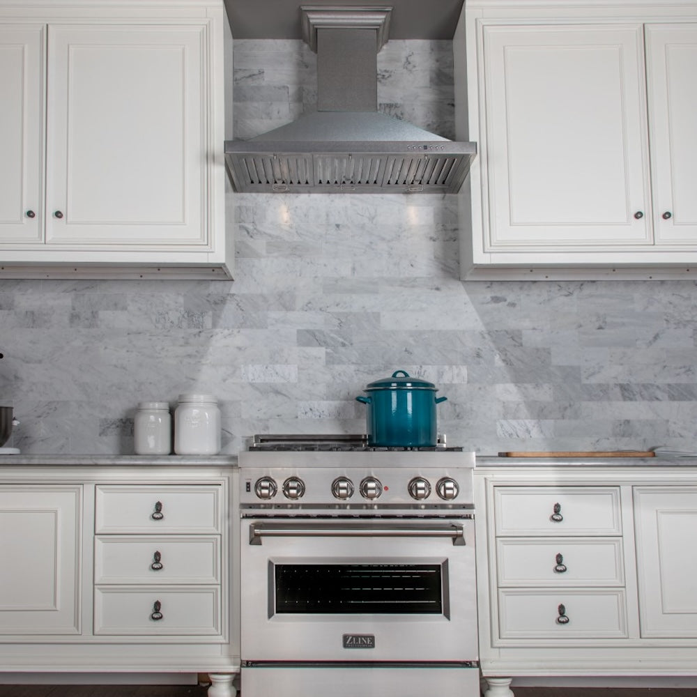 ZLINE Designer Series Satin Stainless Steel Convertible Vent Wall Mount Range Hood (8KBS) in a kitchen with white cabinets, marble backsplash, and a blue pot on the range.