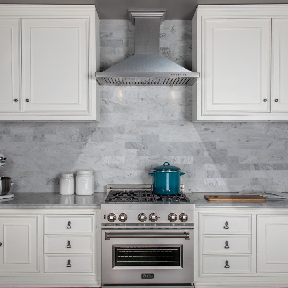ZLINE Designer Series Satin Stainless Steel Convertible Vent Wall Mount Range Hood (8KBS) in a kitchen with white cabinets, marble backsplash, and a blue pot on the range.