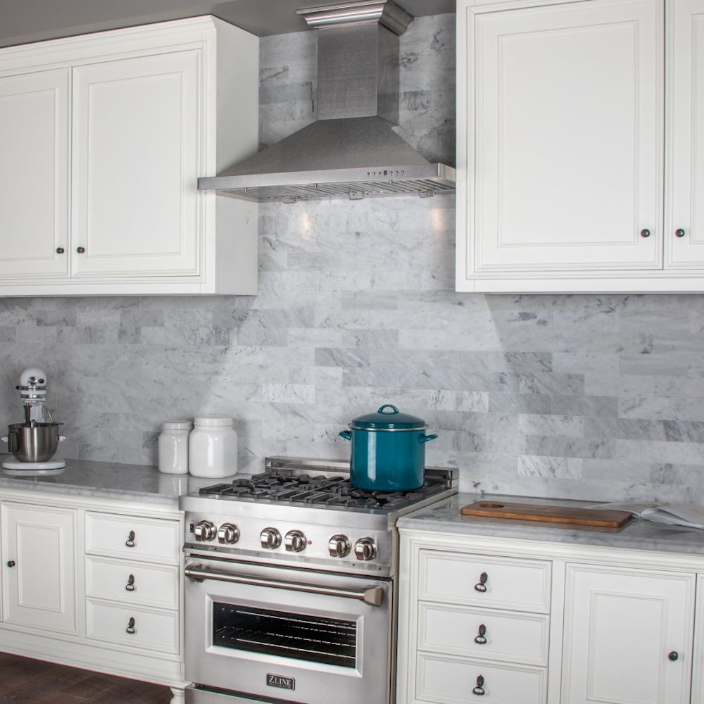 ZLINE Designer Series Satin Stainless Steel Convertible Vent Wall Mount Range Hood (8KBS) in a kitchen with white cabinets, marble backsplash, and a blue pot on the range.