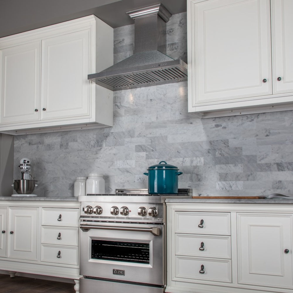 ZLINE Designer Series Satin Stainless Steel Convertible Vent Wall Mount Range Hood (8KBS) in a kitchen with white cabinets, marble backsplash, and a blue pot on the range.