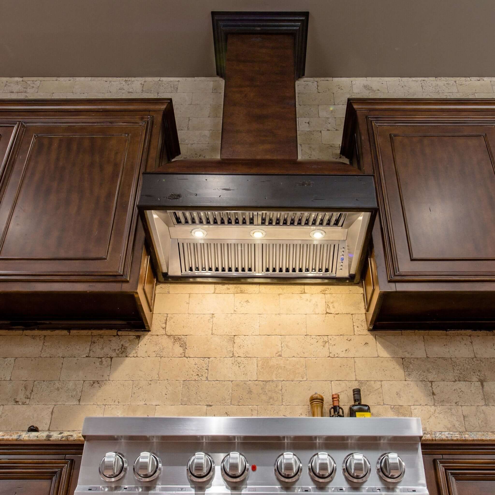 ZLINE 36 in. Wooden Wall Mount Range Hood in Antigua and Walnut (369AW-36) in a rustic kitchen with LED lighting activated and illuminating wall and cooktop.